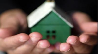 A small, green, wooden home with a white roof, rests in the palms of a person's hand